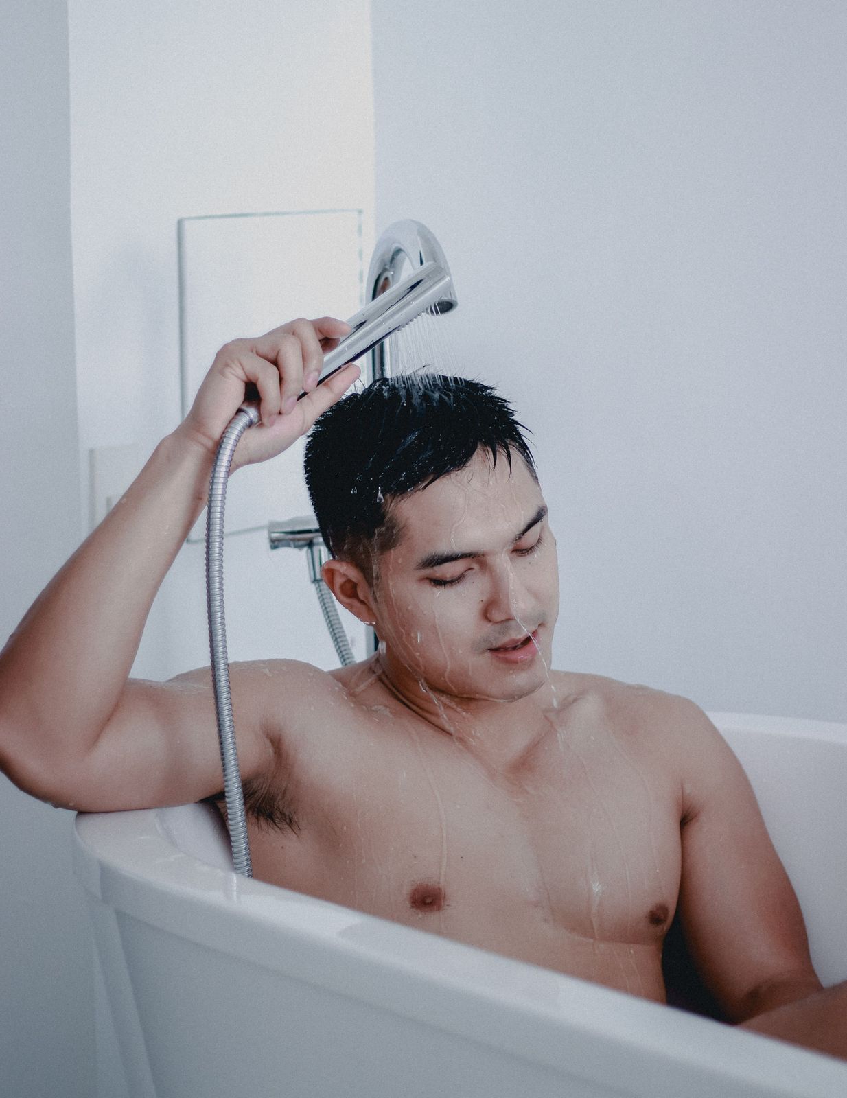 Person seated in a white bathtub rinsing their hair with a slim chrome handheld shower