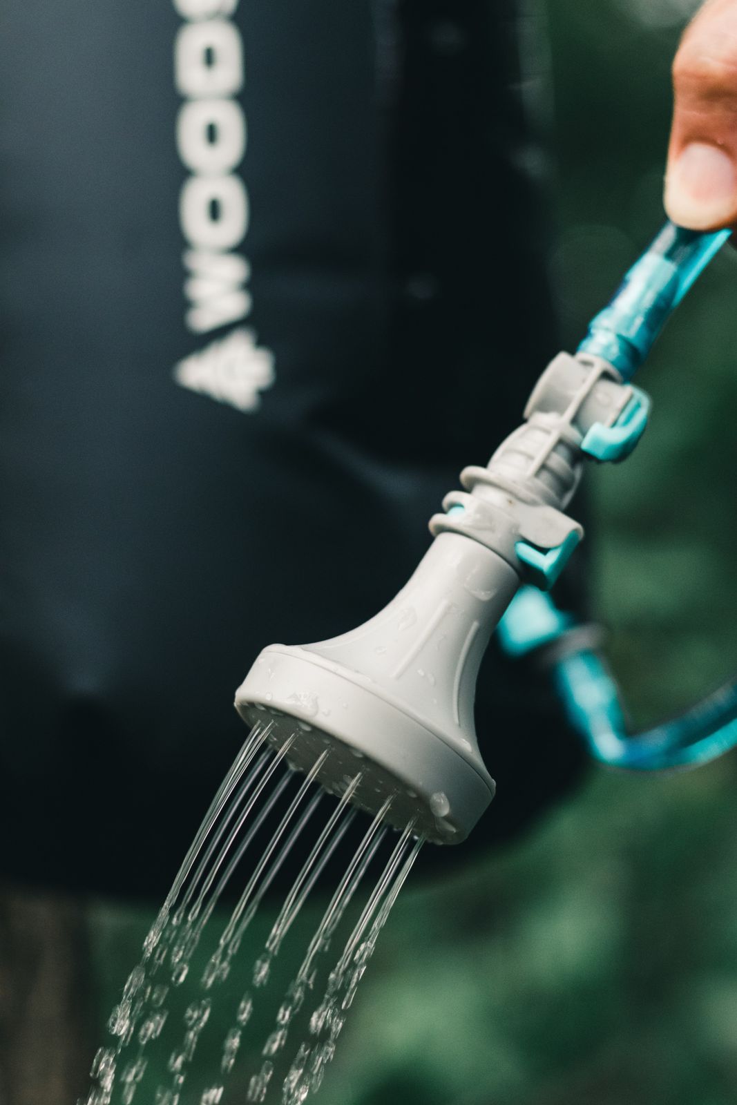Grey outdoor camp shower head with blue tubing releasing streams of water in a green outdoor setting